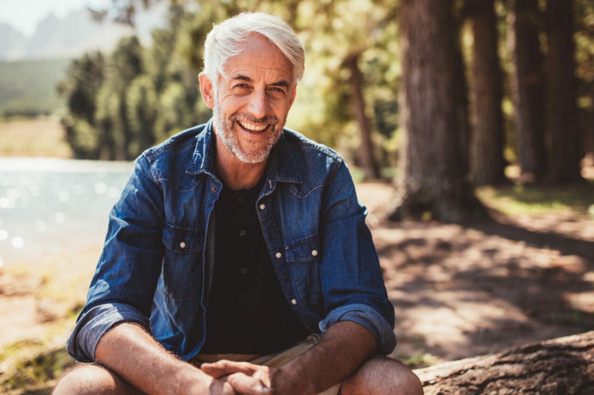 Portrait of happy mature man sitting near a lake looking at camera and smiling. Senior caucasian man sitting on a log by the lake on a summer day.